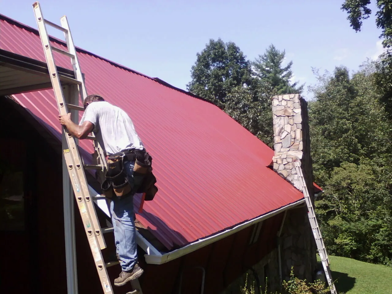 Roofer on a ladder beside a red metal roof and stone chimney — Soffit & Fascia Repair by Roofing Brothers in Acworth